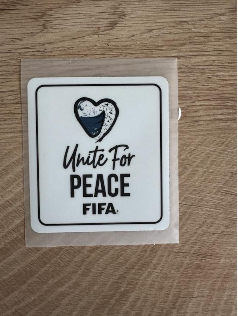 Peut inclure: Un autocollant carr&eacute; blanc aux coins arrondis avec un contour noir. Le motif comprend un c&oelig;ur avec un motif textur&eacute; bleu et blanc, suivi du texte noir Unite For PEACE et du mot FIFA &eacute;crit dans une police de caract&egrave;res plus petite.