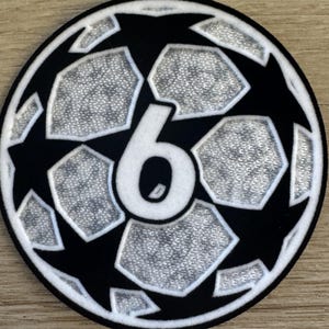 May include: A circular embroidered patch featuring a black and silver soccer ball pattern with a prominent white number 6 in the center. The design includes alternating black star shapes and silver textured hexagonal panels with a thick black border.