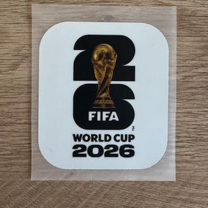 Peut inclure: Un autocollant blanc carr&eacute; aux coins arrondis pr&eacute;sentant le texte noir 26, avec le troph&eacute;e dor&eacute; de la Coupe du Monde de la FIFA plac&eacute; sur le chiffre 2. Sous le graphique, le texte indique FIFA WORLD CUP 2026 en lettres capitales noires.
