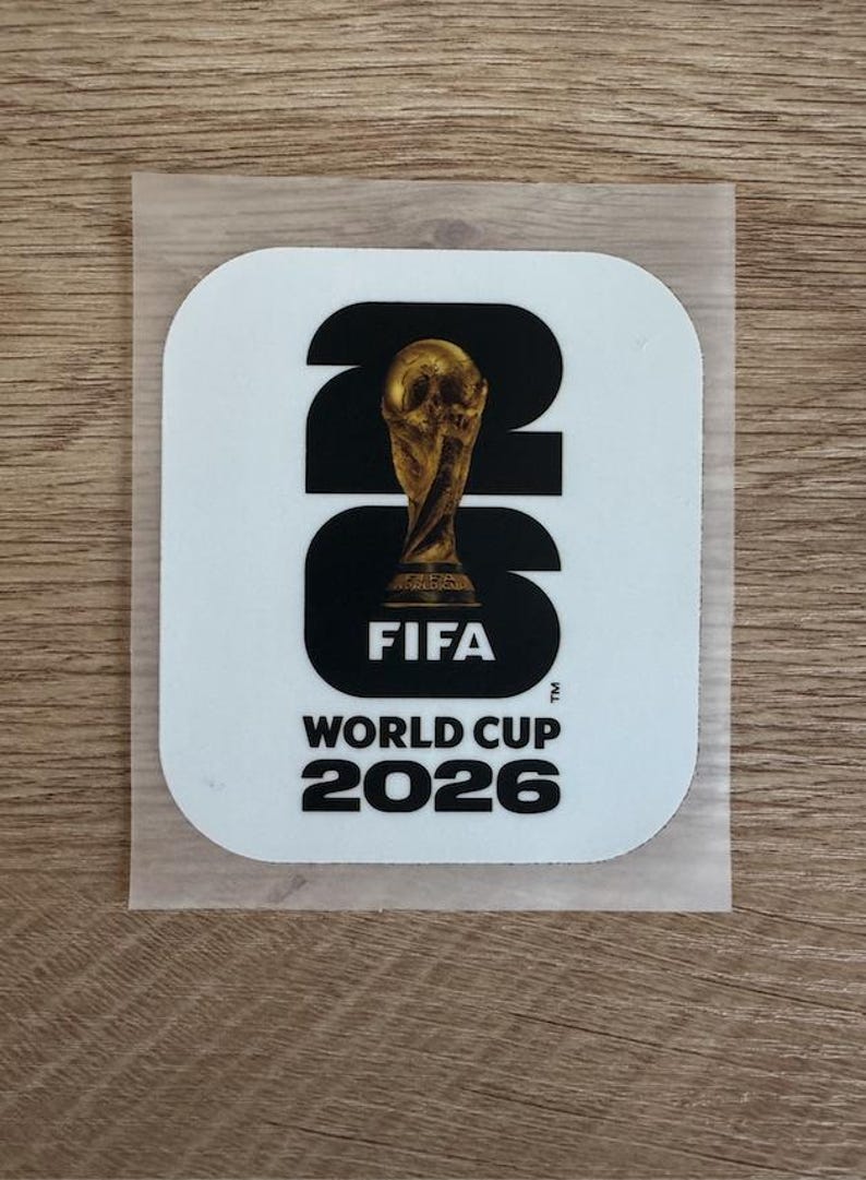 Peut inclure: Un autocollant blanc carr&eacute; aux coins arrondis pr&eacute;sentant le texte noir 26, avec le troph&eacute;e dor&eacute; de la Coupe du Monde de la FIFA plac&eacute; sur le chiffre 2. Sous le graphique, le texte indique FIFA WORLD CUP 2026 en lettres capitales noires.