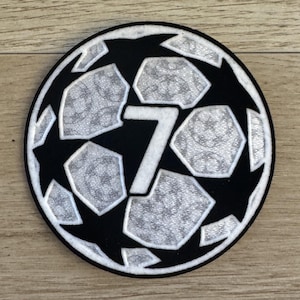 May include: A circular embroidered patch featuring a black and silver soccer ball pattern with a prominent white number 7 in the center. The design includes alternating black star shapes and silver hexagonal panels with a textured, repeating pattern.