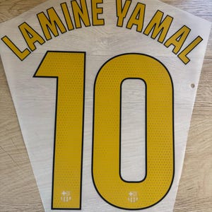May include: A white sports jersey name and number set featuring the name LAMINE YAMAL in yellow letters with black outlines, positioned above the large yellow number 10, which also has black outlines and a small FC Barcelona crest at the base.