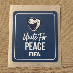 Peut inclure: Un autocollant bleu rectangulaire aux coins arrondis avec un contour de c&oelig;ur blanc contenant une forme bleu fonc&eacute;. Le texte indique Unite For PEACE et FIFA en lettres capitales blanches. L'autocollant a une fine bordure blanche et un support translucide.