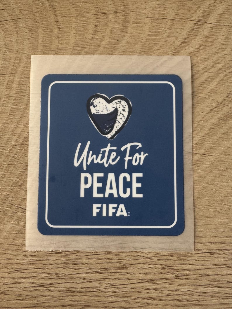 Peut inclure: Un autocollant bleu rectangulaire aux coins arrondis avec un contour de c&oelig;ur blanc contenant une forme bleu fonc&eacute;. Le texte indique Unite For PEACE et FIFA en lettres capitales blanches. L'autocollant a une fine bordure blanche et un support translucide.