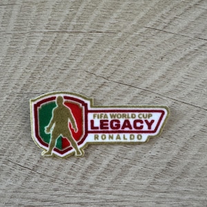 Peut inclure: Un &eacute;cusson brod&eacute; rectangulaire pr&eacute;sentant un bouclier sur la gauche avec la silhouette dor&eacute;e d'un joueur de football sur un fond vert et rouge. &Agrave; droite, le texte indique FIFA WORLD CUP LEGACY RONALDO en lettres rouges et dor&eacute;es.