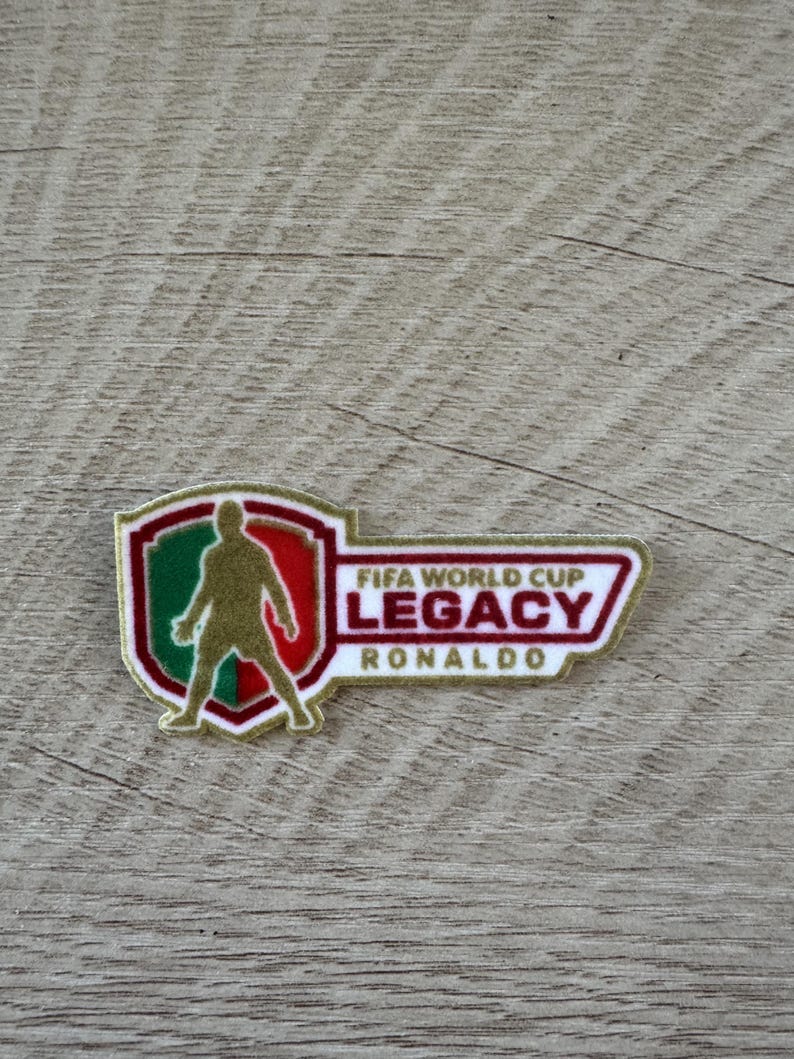 Peut inclure: Un &eacute;cusson brod&eacute; rectangulaire pr&eacute;sentant un bouclier sur la gauche avec la silhouette dor&eacute;e d'un joueur de football sur un fond vert et rouge. &Agrave; droite, le texte indique FIFA WORLD CUP LEGACY RONALDO en lettres rouges et dor&eacute;es.