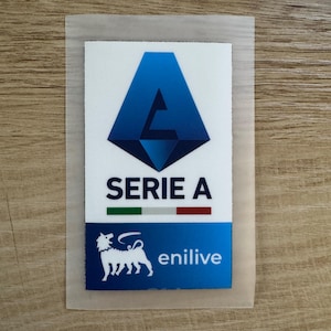 May include: A rectangular patch featuring the blue Serie A logo at the top, the text SERIE A, a small green, white, and red horizontal stripe, and a blue bottom section with a white six-legged dog icon and the white text enilive.