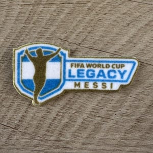 May include: A rectangular patch with a shield-shaped left side featuring a gold silhouette of a person with arms raised over a blue and white striped background. The right side contains the text FIFA WORLD CUP LEGACY MESSI in blue and gold.