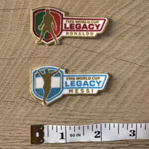 Peut inclure: Deux &eacute;cussons brod&eacute;s avec des motifs FIFA World Cup Legacy. L'&eacute;cusson du haut montre une silhouette dor&eacute;e de Ronaldo sur un bouclier vert, blanc et rouge. Celui du bas montre une silhouette dor&eacute;e de Messi sur un bouclier bleu et blanc. Un ruban &agrave; mesurer indique une largeur d'environ 7,6 cm.