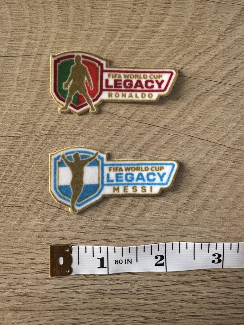 Peut inclure: Deux &eacute;cussons brod&eacute;s avec des motifs FIFA World Cup Legacy. L'&eacute;cusson du haut montre une silhouette dor&eacute;e de Ronaldo sur un bouclier vert, blanc et rouge. Celui du bas montre une silhouette dor&eacute;e de Messi sur un bouclier bleu et blanc. Un ruban &agrave; mesurer indique une largeur d'environ 7,6 cm.