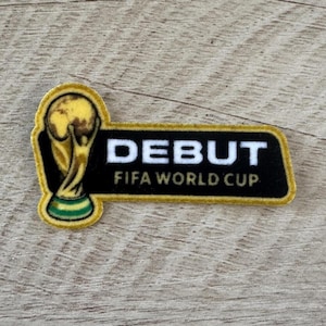 May include: A rectangular embroidered patch with rounded edges featuring a gold and green FIFA World Cup trophy icon on the left and the white text DEBUT above the gold text FIFA WORLD CUP on a black background with a gold border.
