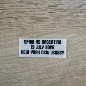 May include: A rectangular white fabric label featuring black embroidered text that reads: SPAIN US ARGENTINA, 19 JULY 2026, NEW YORK NEW JERSEY. The label has a clean, straight-cut edge and is designed for attachment to garments or other textile items.