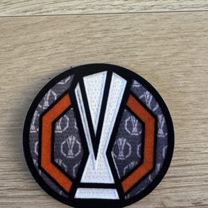 May include: A circular embroidered patch featuring a central white trophy icon outlined in black. The design includes two orange hexagonal shapes flanking the trophy, set against a dark gray background patterned with repeating trophy silhouettes.