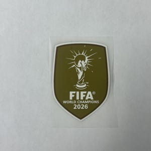 May include: A shield-shaped patch in olive green with a white border. The center features a white graphic of the FIFA World Cup trophy with radiating lines. Below the trophy, the text reads FIFA WORLD CHAMPIONS 2026 in white capital letters.