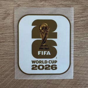 Peut inclure: Un autocollant carr&eacute; aux coins arrondis avec un fond blanc et une bordure dor&eacute;e. Le design pr&eacute;sente les grands chiffres dor&eacute;s 26 avec le troph&eacute;e de la Coupe du Monde de la FIFA centr&eacute; dans le z&eacute;ro, suivi du texte FIFA WORLD CUP 2026.