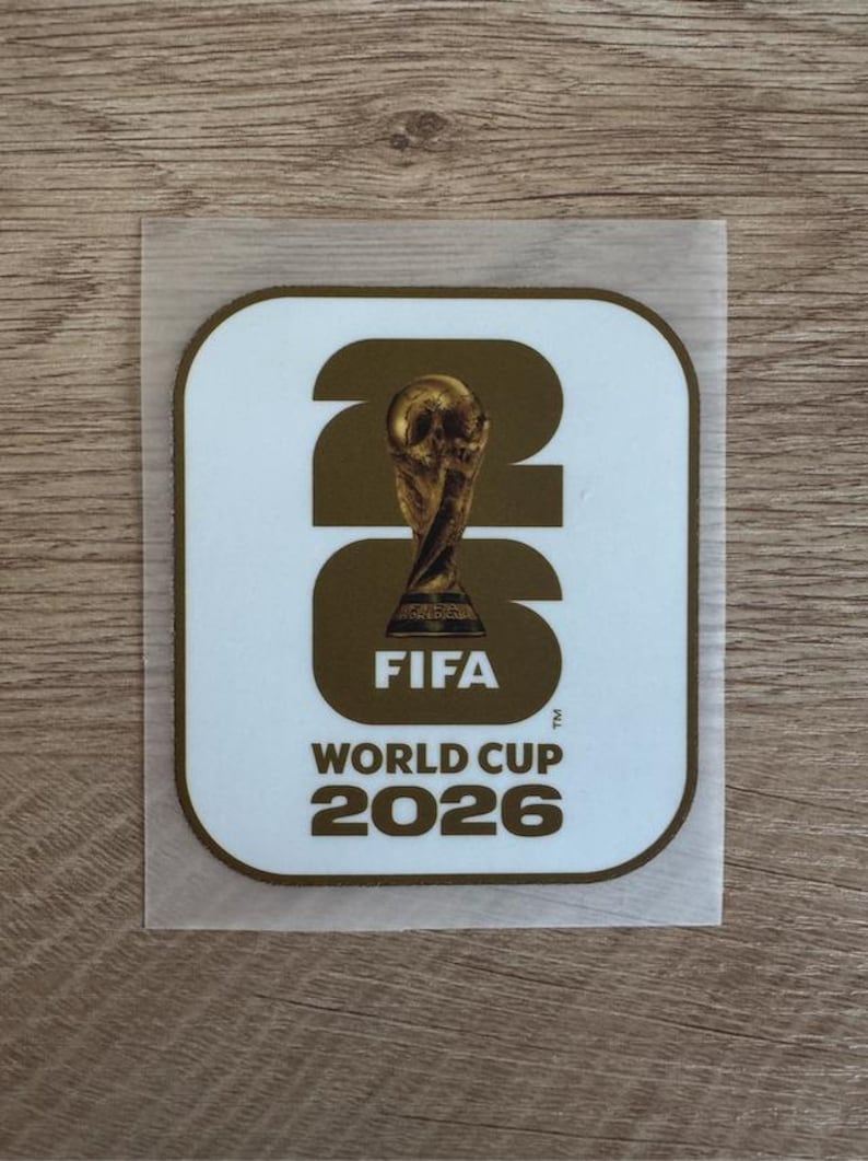 Peut inclure: Un autocollant carr&eacute; aux coins arrondis avec un fond blanc et une bordure dor&eacute;e. Le design pr&eacute;sente les grands chiffres dor&eacute;s 26 avec le troph&eacute;e de la Coupe du Monde de la FIFA centr&eacute; dans le z&eacute;ro, suivi du texte FIFA WORLD CUP 2026.