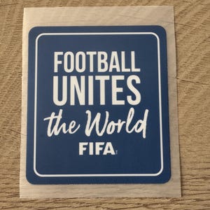 Peut inclure: Un autocollant rectangulaire bleu aux coins arrondis avec le texte blanc FOOTBALL UNITES the World et le logo FIFA. L'autocollant pr&eacute;sente une fine bordure blanche et est pr&eacute;sent&eacute; sur un support transparent avec un bord adh&eacute;sif visible.