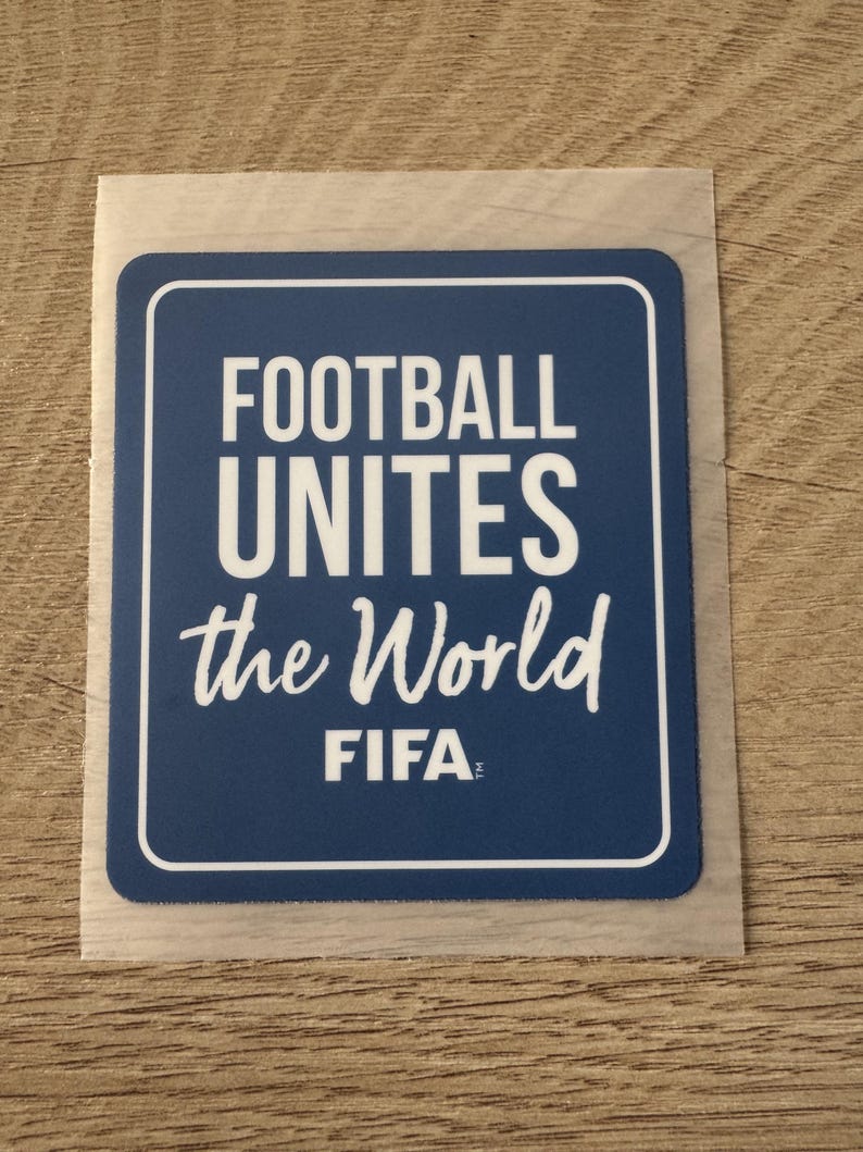 Peut inclure: Un autocollant rectangulaire bleu aux coins arrondis avec le texte blanc FOOTBALL UNITES the World et le logo FIFA. L'autocollant pr&eacute;sente une fine bordure blanche et est pr&eacute;sent&eacute; sur un support transparent avec un bord adh&eacute;sif visible.