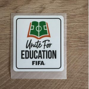 Peut inclure: Un autocollant carr&eacute; blanc aux coins arrondis pr&eacute;sentant un graphique vert et orange d'un livre ouvert en forme de terrain de football. Sous le graphique, le texte indique Unite For EDUCATION FIFA en lettres capitales noires, stylis&eacute;es et grasses.