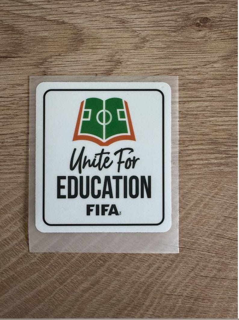 Peut inclure: Un autocollant carr&eacute; blanc aux coins arrondis pr&eacute;sentant un graphique vert et orange d'un livre ouvert en forme de terrain de football. Sous le graphique, le texte indique Unite For EDUCATION FIFA en lettres capitales noires, stylis&eacute;es et grasses.