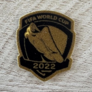 May include: A shield-shaped patch featuring a black and gold color scheme. The text FIFA WORLD CUP is arched at the top, with the year 2022 printed at the bottom. A central gold graphic depicts a stylized soccer trophy silhouette on a black background.