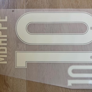 May include: A white heat transfer decal sheet featuring the name MBAPPÉ in vertical block letters and the number 10 in two different sizes. Each number includes a small circular crest detail. The design is printed on a clear, semi-transparent film.