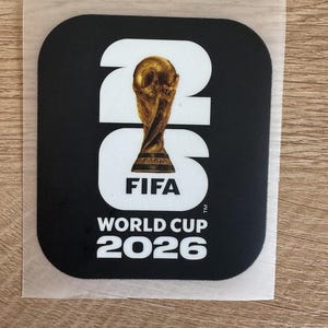 Peut inclure: Un autocollant noir carr&eacute; aux coins arrondis pr&eacute;sentant le texte blanc 26, avec le troph&eacute;e dor&eacute; de la Coupe du Monde de la FIFA rempla&ccedil;ant le chiffre 0. En dessous, le texte indique FIFA WORLD CUP 2026 en lettres capitales blanches.