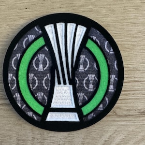 May include: A circular embroidered patch featuring a central white trophy icon outlined in black. The background consists of a dark gray fabric with a repeating white trophy pattern, framed by a bright green circular border and a solid black outer edge.