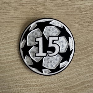 May include: A circular patch featuring a black and silver soccer ball pattern with the number 15 prominently displayed in the center. The design includes alternating black star shapes and silver hexagonal panels, all enclosed within a thick black border.