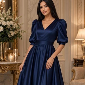 May include: A navy blue satin dress featuring a V-neckline, a fitted waist, and elbow-length puff sleeves. The skirt is full and gathered at the waist, creating a flowing silhouette. The fabric has a smooth, lustrous finish with a subtle sheen.