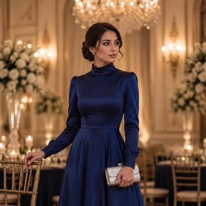 May include: A long-sleeved navy blue satin dress featuring a high mock neckline, fitted bodice, and a full skirt. The dress has cuffed sleeves and is paired with a rectangular, metallic silver clutch held in one hand.