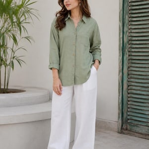 May include: A long-sleeved sage green button-down shirt with a collar and a single chest pocket, paired with loose-fitting white wide-leg trousers. The shirt features light-colored buttons down the front and is worn with simple beige open-toe sandals.