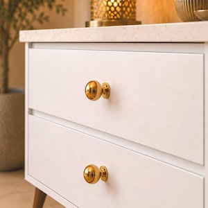 May include: A white two-drawer chest featuring two polished gold spherical knobs on the front. The chest has a light-colored stone top and is supported by tapered wooden legs. The text Maison Artisanat Kech is visible in the bottom right corner.