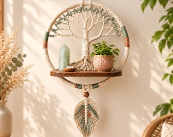 May include: A circular macrame wall hanging featuring a tree design in cream, sage green, and brown cord. It includes a dark wooden shelf holding a green crystal point and a small potted plant. A single macrame feather hangs from the bottom.