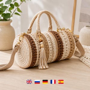 May include: A cylindrical crochet handbag featuring alternating horizontal stripes in cream, beige, and dark brown. The bag includes two top handles, a detachable shoulder strap with gold-tone hardware, and a decorative tassel charm with wooden beads.