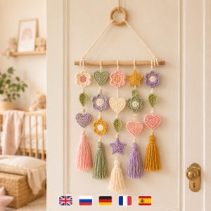 Peut inclure: Une décoration murale composée d'une tige en bois horizontale suspendue par un anneau en bois. Cinq brins verticaux de formes crochetées, incluant des fleurs, cœurs, étoiles et feuilles aux tons pastel, se terminent par des pompons en laine.