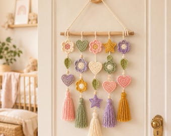 May include: A wall hanging featuring a horizontal wooden rod suspended by a wooden ring. Five vertical strands of crocheted shapes including flowers, hearts, stars, and leaves in pastel colors are attached, each ending with a yarn tassel.