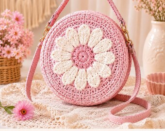 May include: A round, pink woven crossbody bag featuring a large, white, crocheted daisy flower design on the front. The bag includes a matching pink shoulder strap attached with gold-toned metal hardware, creating a structured, circular silhouette.