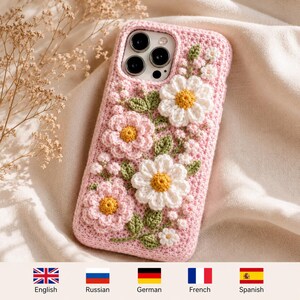 May include: A light pink crochet phone case featuring three-dimensional white and pink flowers with yellow centers and green leaves. The case has a textured, hand-stitched appearance and covers the back and sides of a smartphone with a triple camera lens.