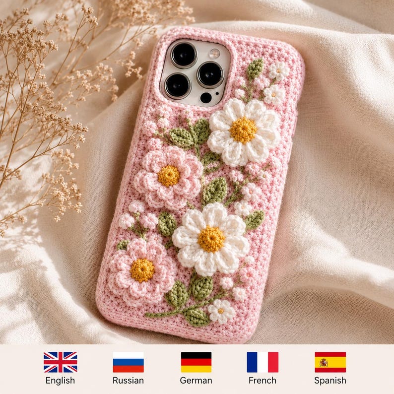 May include: A light pink crochet phone case featuring three-dimensional white and pink flowers with yellow centers and green leaves. The case has a textured, hand-stitched appearance and covers the back and sides of a smartphone with a triple camera lens.