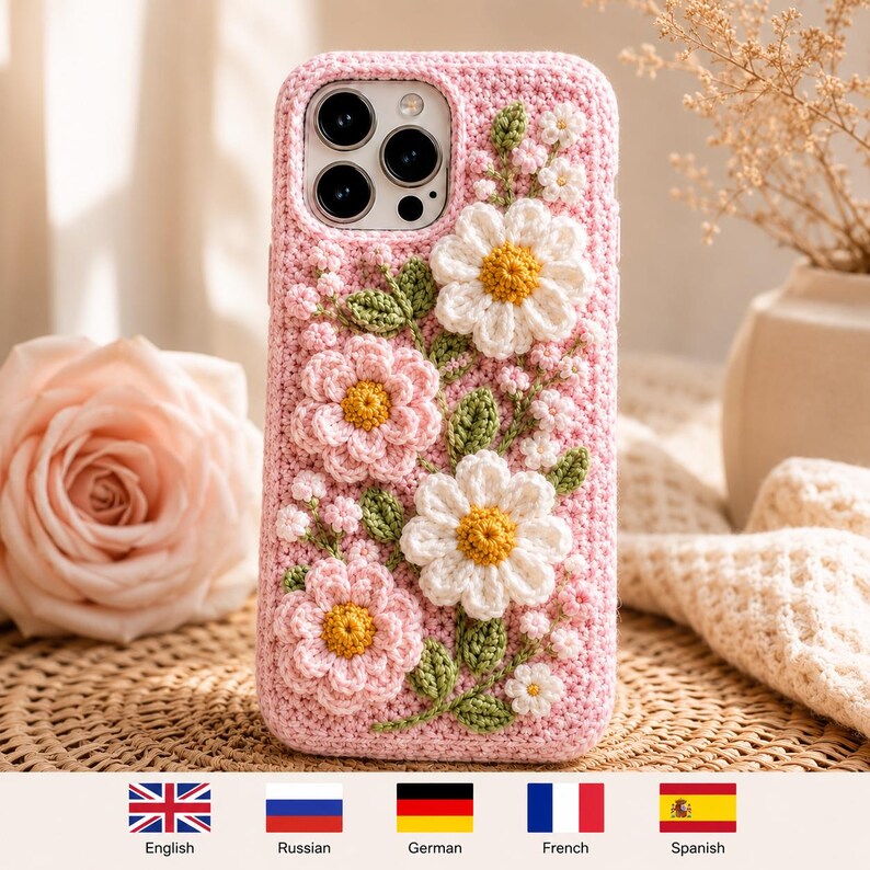 May include: A light pink crochet phone case featuring three-dimensional floral embellishments. The design includes two large white daisies with yellow centers, two pink flowers with yellow centers, small white buds, and green crocheted leaves.