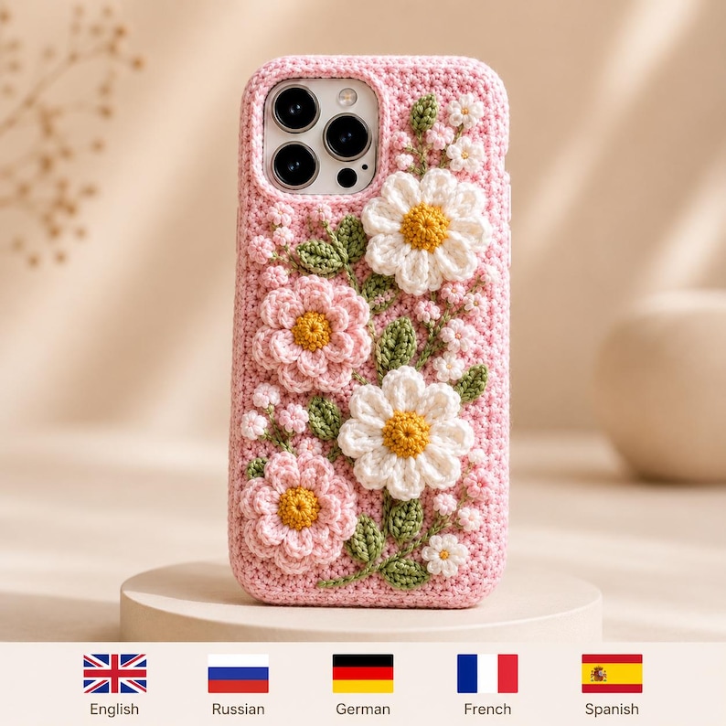 May include: A light pink crochet phone case featuring three-dimensional white and pink flowers with yellow centers and green leaves. The case has a textured, hand-stitched appearance and covers the back and sides of a smartphone with a triple camera.