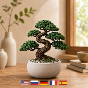 Könnte beinhalten: Ein gehäkelter Bonsai-Baum mit einem strukturierten braunen Stamm und leuchtend grünem Blattwerk, eingetopft in einen runden, hellgrauen Keramikbehälter, der an der Basis mit kleinen, glatten, mehrfarbigen Kieselsteinen gefüllt ist.