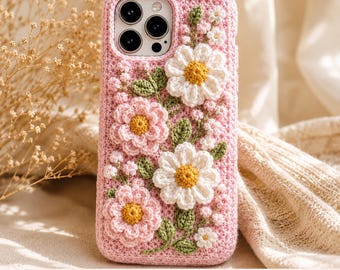 Puede incluir: Una funda de teléfono de ganchillo rosa con adornos florales tridimensionales. El diseño incluye margaritas blancas con centros amarillos, flores rosas con centros amarillos y pequeñas hojas y tallos verdes sobre la superficie texturizada.