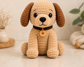 Puede incluir: Un juguete de peluche tejido a ganchillo con forma de perro, con pelaje color canela claro, orejas marrón oscuro y un collar marrón oscuro. El perro tiene ojos de plástico negro, nariz bordada en negro y una etiqueta redonda con un corazón.
