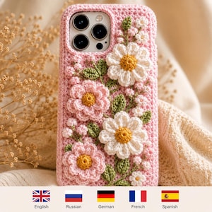 May include: A pink crochet phone case featuring three-dimensional floral embellishments. The design includes white and pink crocheted flowers with yellow centers, accompanied by green crocheted leaves and small buds, all integrated into a textured pink base.