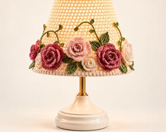 May include: A table lamp featuring a cream-colored crocheted lampshade adorned with three-dimensional fabric roses in shades of pink, dark rose, and white, accented with green crocheted leaves and vines, set on a smooth, glossy cream-colored base.