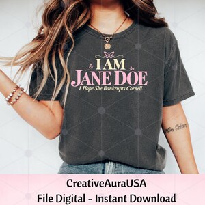 May include: A charcoal grey short-sleeve t-shirt featuring the text I AM JANE DOE I Hope She Bankrupts Cornell in yellow and pink lettering. The design includes small pink butterfly graphics above the text and a crew neckline.