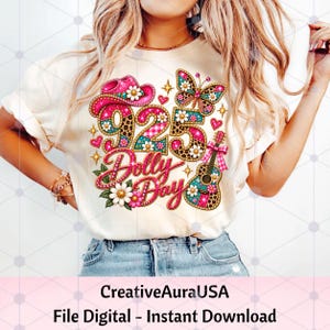 May include: A cream-colored t-shirt featuring a graphic design with the text 925 Dolly Day. The design includes a pink cowboy hat, a butterfly, a guitar, and daisies, all decorated with leopard print, pink gingham, and bright pink accents.
