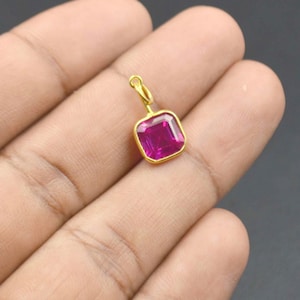 Puede incluir: Un pequeño colgante con una piedra preciosa de color magenta intenso de corte cuadrado, engastada en un marco de metal dorado pulido. El colgante incluye una pequeña anilla circular dorada en la parte superior para una cadena.